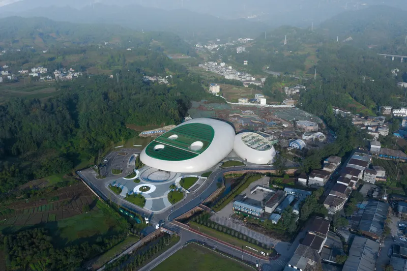 Aerial wide: the full site with both volumes, green roofs, circular forecourt plaza, surrounding town, misty forested hills