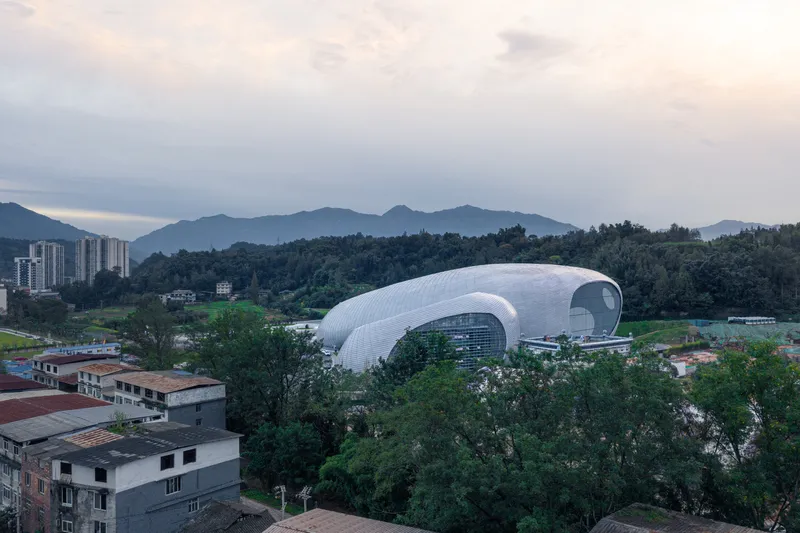 From the town at dusk: the larger volume rising above the rooftops, curved white aluminium skin, forested hillside, mountains in haze