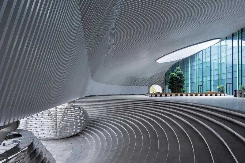Sunken entry plaza: ribbed aluminium walls curving upward, tiered concrete steps descending to the lobby, oval skylights above, trees visible through the glazing