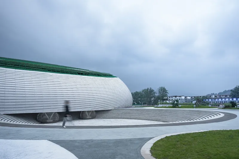Side elevation: horizontal aluminium ribs wrapping the curved form, sunken entry visible at left, green roof edge, figure passing, overcast