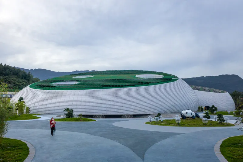 Approach: curved white aluminium facade with horizontal ribbing, green roof visible above, panda sculptures at the entrance, figure walking, overcast sky