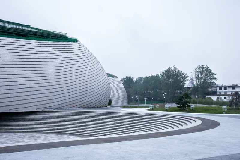 Sunken amphitheatre: tiered concrete steps descending toward the building base, ribbed aluminium skin rising above, green roof edge, trees beyond
