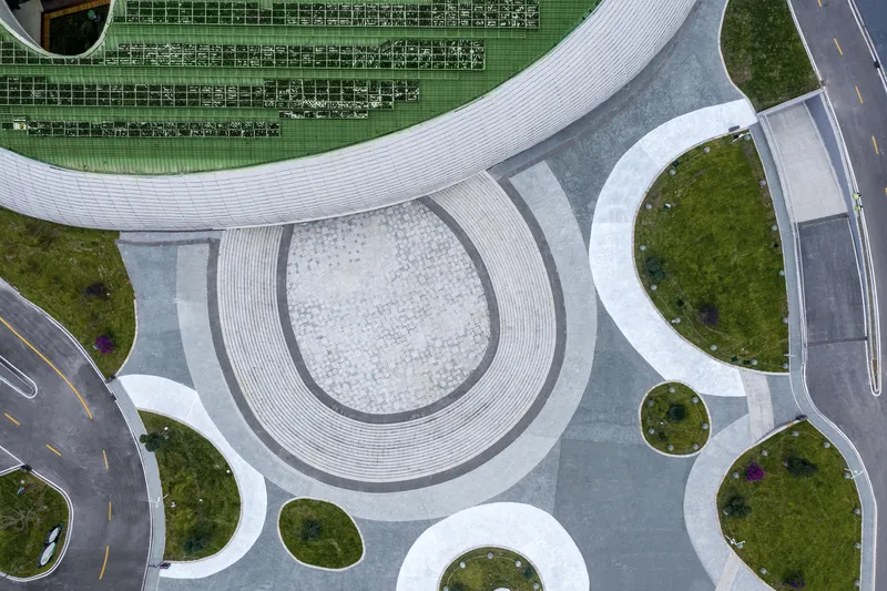 Aerial detail: green roof with rows of solar panels, sunken entry plaza below with patterned paving, circular landscape beds, roads curving around the building