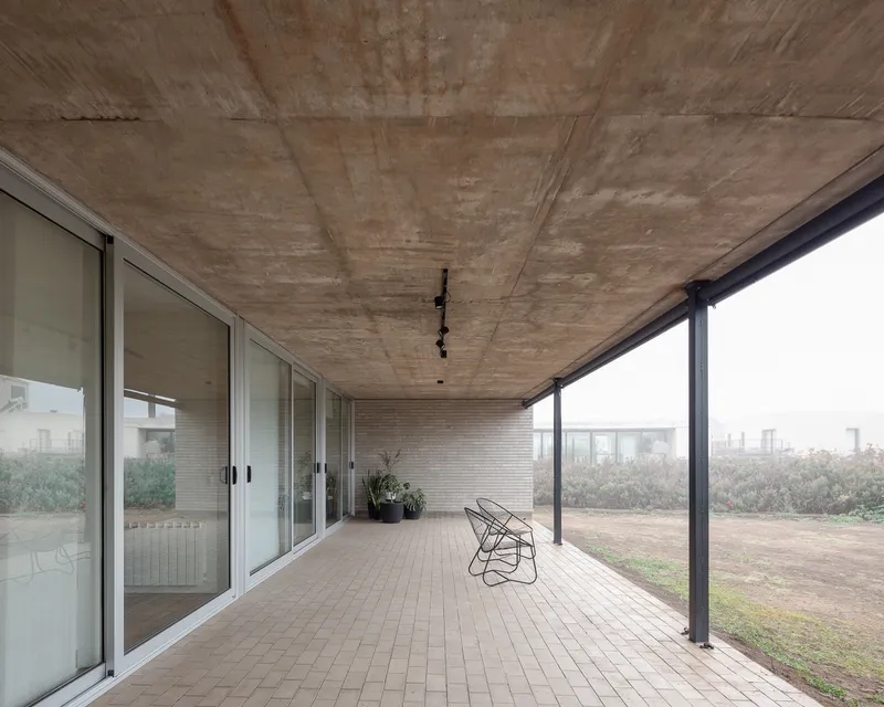 Covered porch: exposed concrete ceiling, brick floor, wire chair, full-height glass to garden