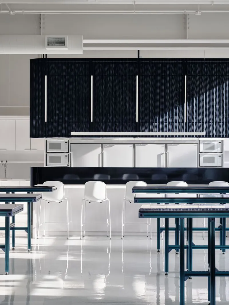 Cafeteria with white stackable chairs, blue metal tables and a dark ribbed island under exposed ceiling