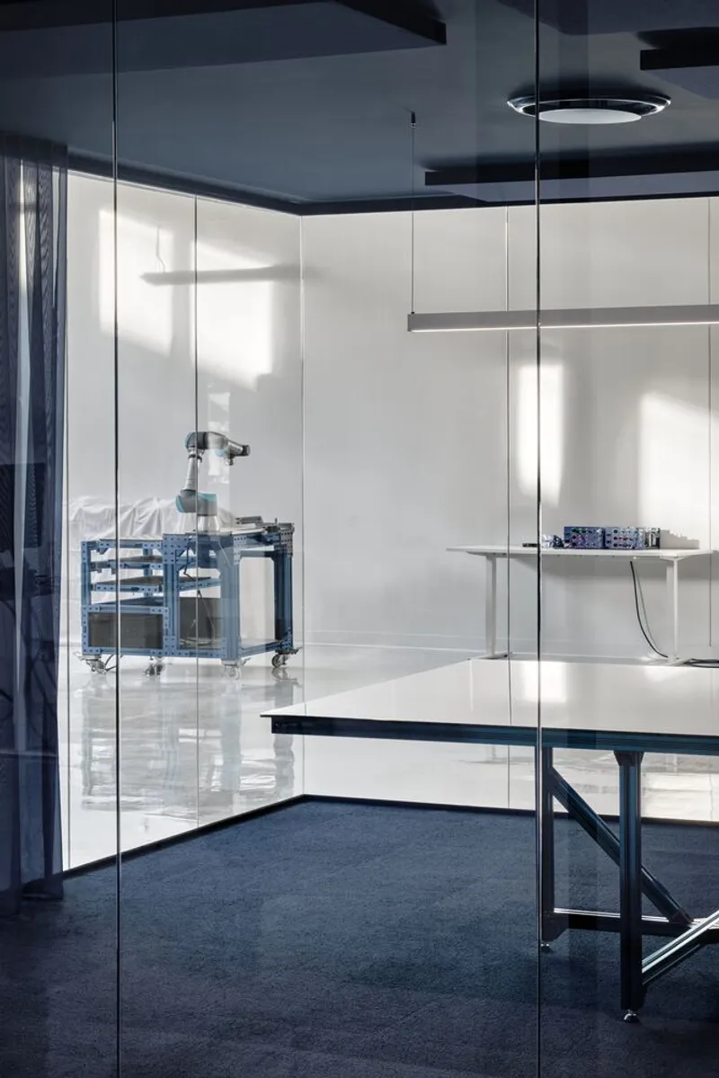 Glass partition dividing a white-walled room from a space with blue carpet and suspended fixtures