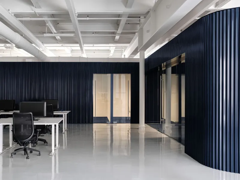 Long view of dark blue corrugated walls with illuminated doorways and reflective white floor under exposed structure