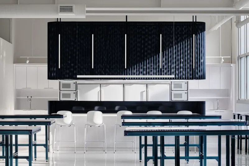 Kitchen area with dark fluted canopy over counters, white stools and workbenches below suspended linear lights