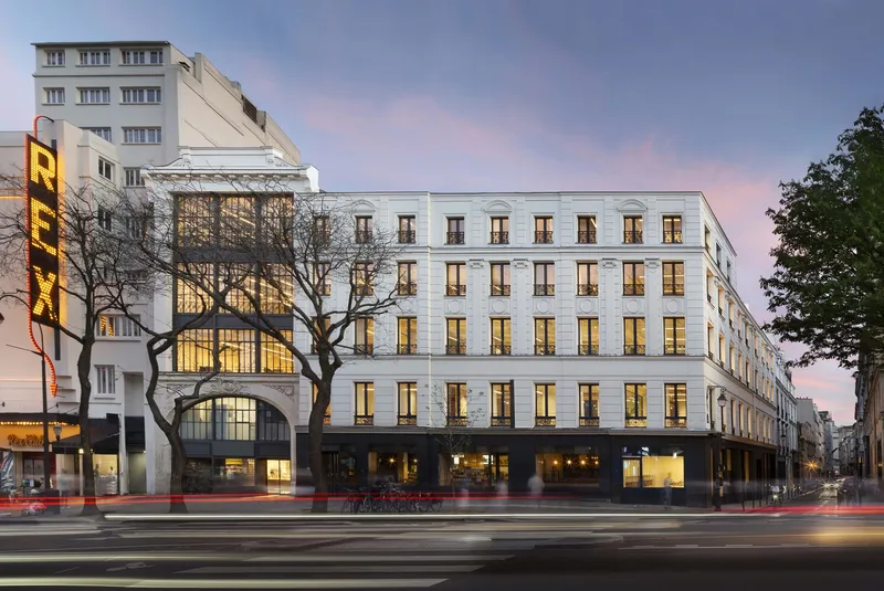 Street facade at dusk: restored Haussmann building with illuminated ground-floor steel and glass windows, Grand Rex cinema next door
