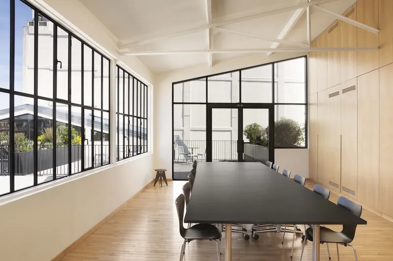 Top-floor meeting room: steel-frame windows, white-painted trusses, long dark table, Parisian rooftops outside