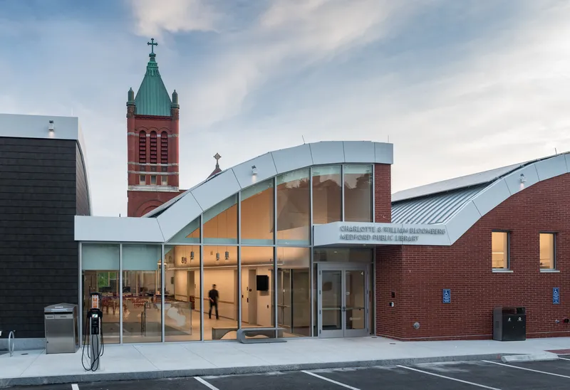 Glass-fronted entrance with folded metal roof connecting brick volumes beneath a copper-clad bell tower