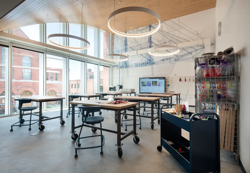 Classroom with timber ceiling, circular pendant lights, and mobile work tables facing floor-to-ceiling windows