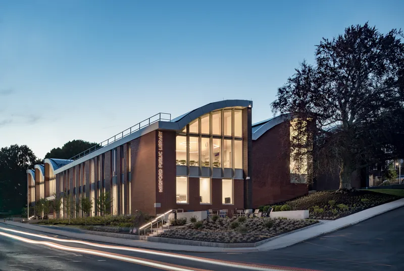 Brick and glass facade with curved roofline illuminated at dusk beside a mature tree