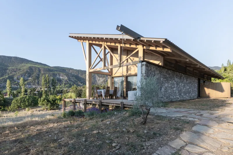 Covered terrace with exposed timber rafters and stone masonry base facing mountain foothills at dusk
