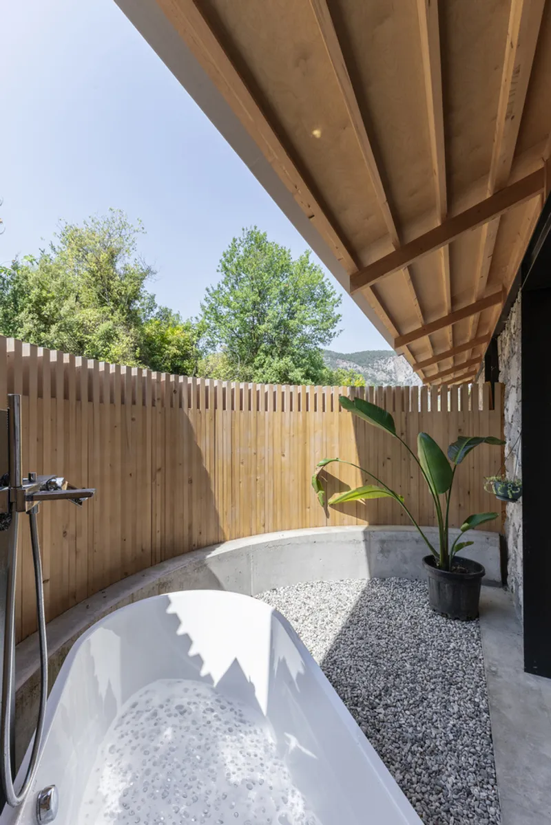 Outdoor bathtub beneath a timber-framed canopy with vertical slatted fence and potted plants in gravel