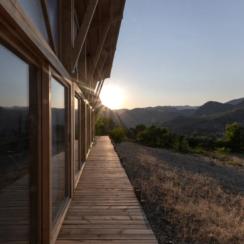 Timber deck walkway along glazed facade with exposed rafters at sunset overlooking mountain landscape