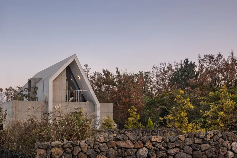 Gabled concrete volume with triangular glass facade rising above a dry stone retaining wall at dusk