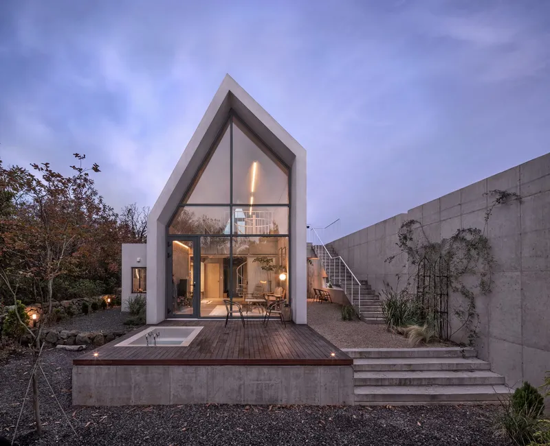 Glazed gable facade and timber deck terrace with concrete stairs ascending the hillside at twilight