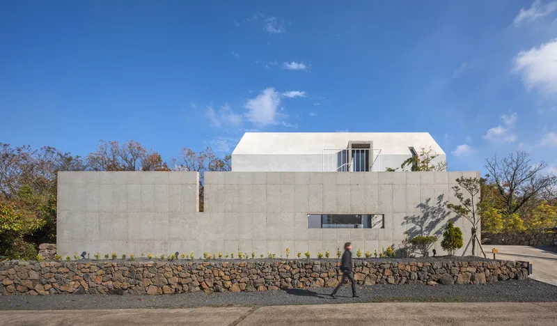 Street view of the concrete and white metal-clad volumes behind a stone retaining wall with a figure walking