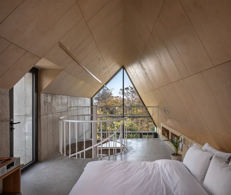 Bedroom with diagonal wood-paneled ceiling and triangular window overlooking treetops in autumn