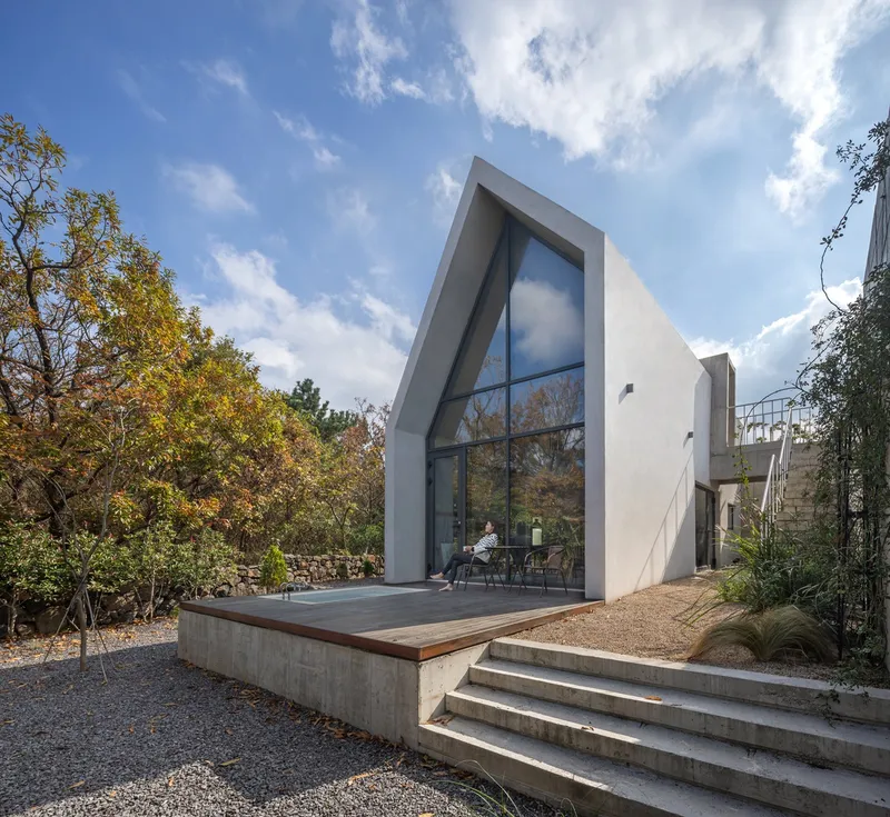 White gabled volume with full-height glazing and timber deck overlooking gravel courtyard and autumn foliage