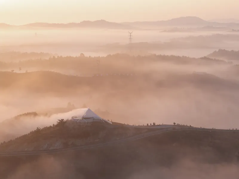 Dawn aerial (repeated): membrane tower on its hilltop emerging from mist, terraces visible below