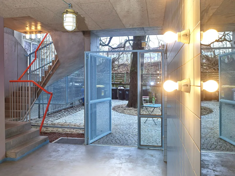 Entry lobby with concrete staircase, red handrail, glass doors opening to cobblestone courtyard with leafless tree