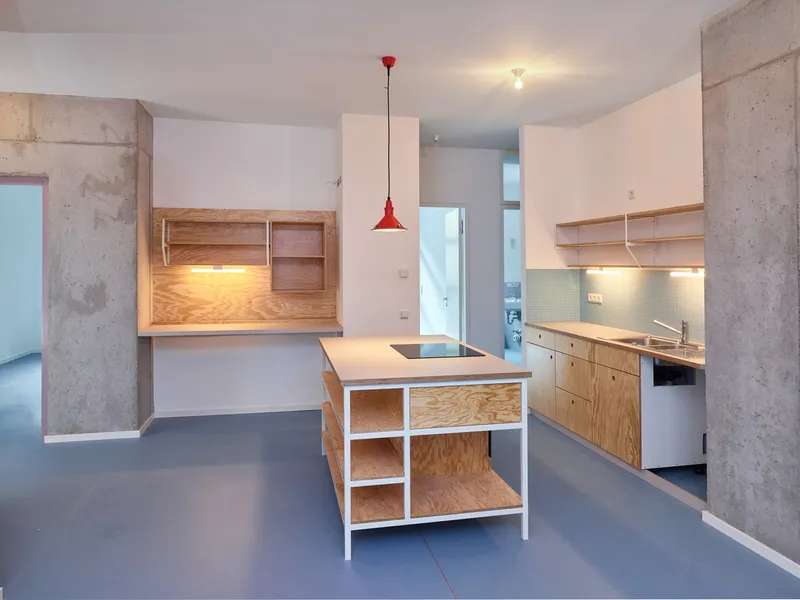 Kitchen with plywood island, white countertops, red pendant light, and exposed concrete columns