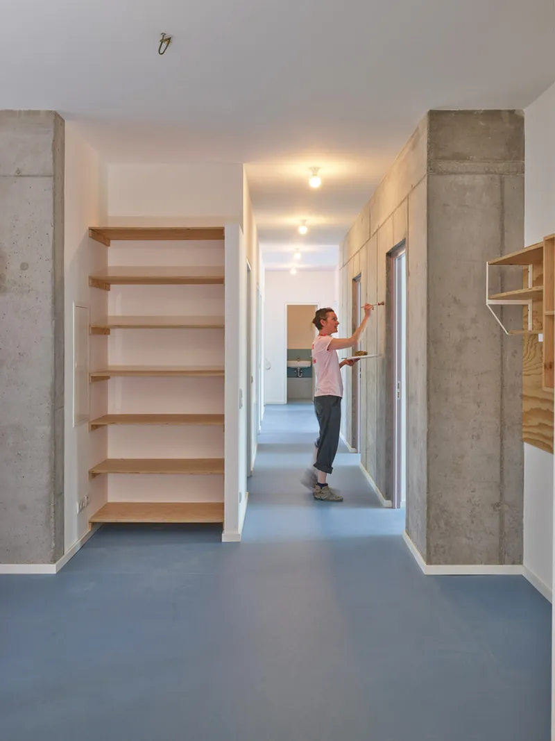 Interior hallway with exposed concrete columns, blue flooring, plywood shelving, and a resident adjusting a door