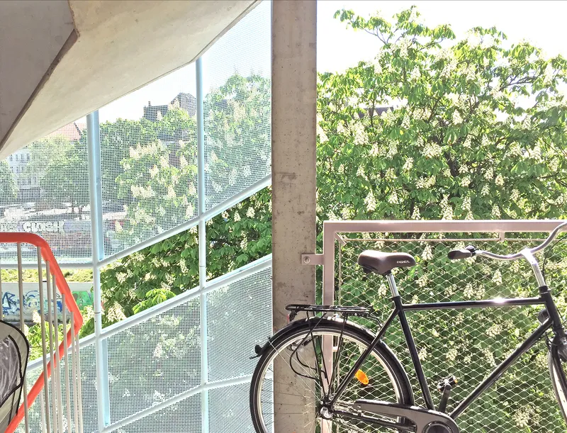 Balcony corner with bicycle, perforated metal railing, concrete column, and flowering tree beyond the mesh screen
