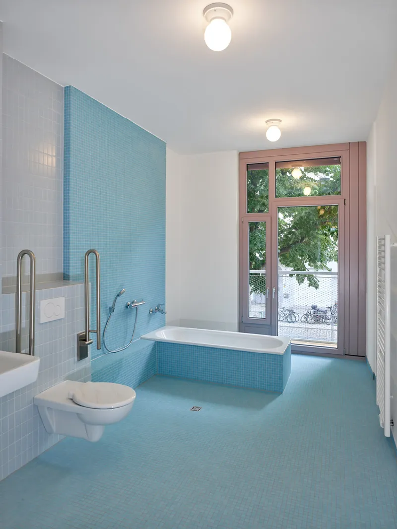 Bathroom with blue mosaic tile, sunken tub, wall-mounted toilet, and pink-framed window overlooking street