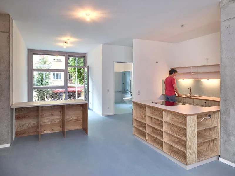 Open kitchen and living area with plywood cabinetry, grey flooring, and exposed concrete columns