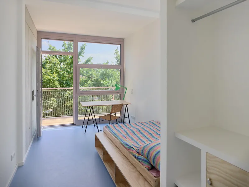 Bedroom with plywood bed frame, blue flooring, and glazed door opening to balcony with trees beyond