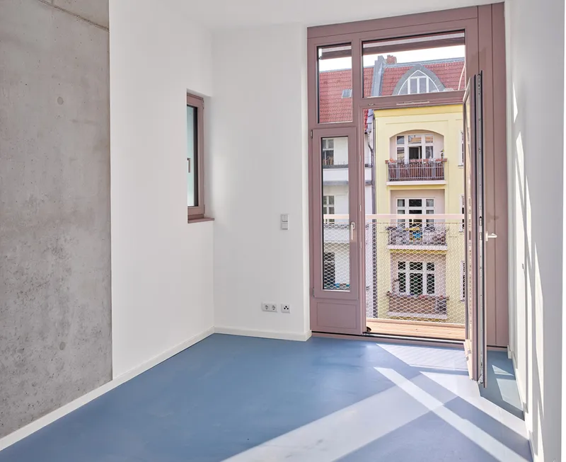 Room with blue flooring, exposed concrete wall, and pink-framed doors opening to balcony with metal mesh railing