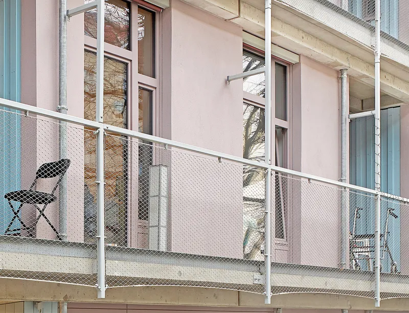 Narrow balcony with white mesh railing, folding chair, and pale pink facade with teal window frames