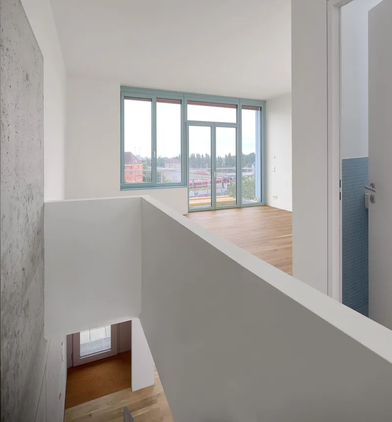 Double-height interior void with white walls, timber flooring, and teal-framed windows overlooking rooftops