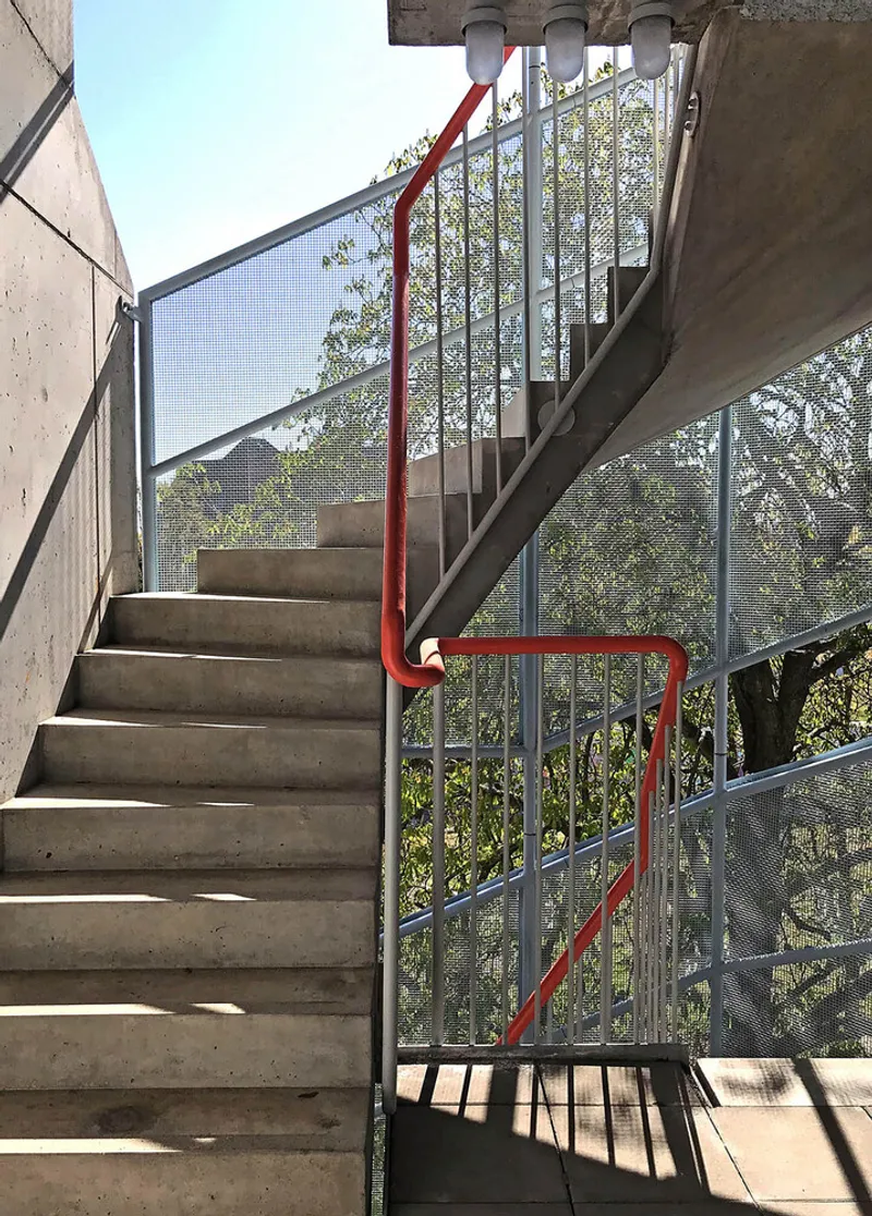 Concrete staircase with red metal handrail and perforated metal balustrade backed by green foliage