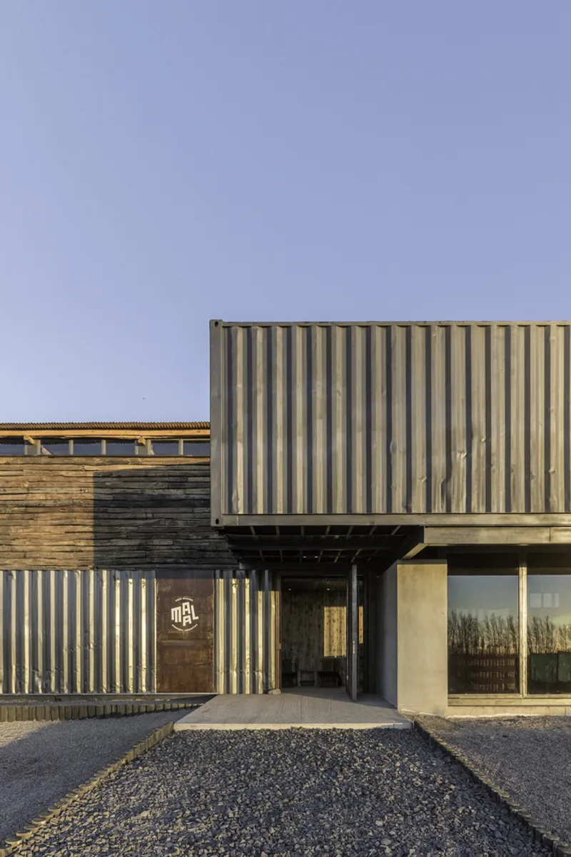 Entry facade composed of corrugated metal panels and timber cladding with a gravel courtyard at dusk