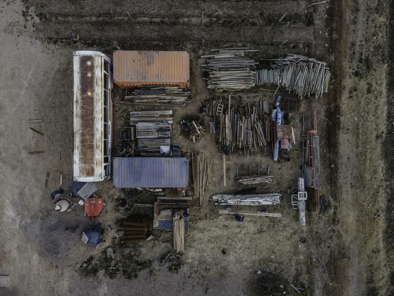 Aerial view of a construction site with shipping containers, stacked timber frames and material piles on gravel
