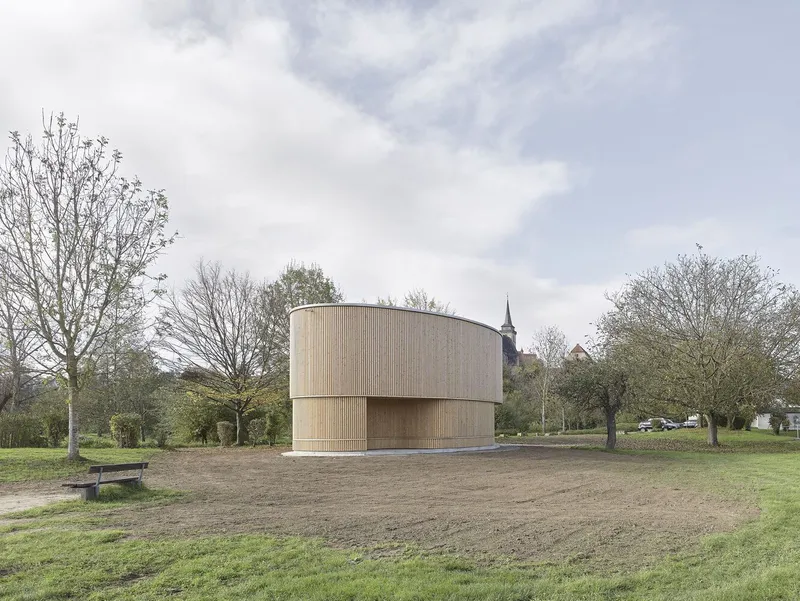 Cylindrical timber pavilion on a park field with church spire visible through bare trees