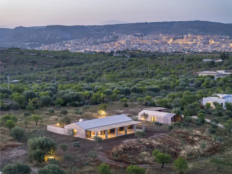 Distant view of the low-lying residence lit from within at dusk, surrounded by olive groves
