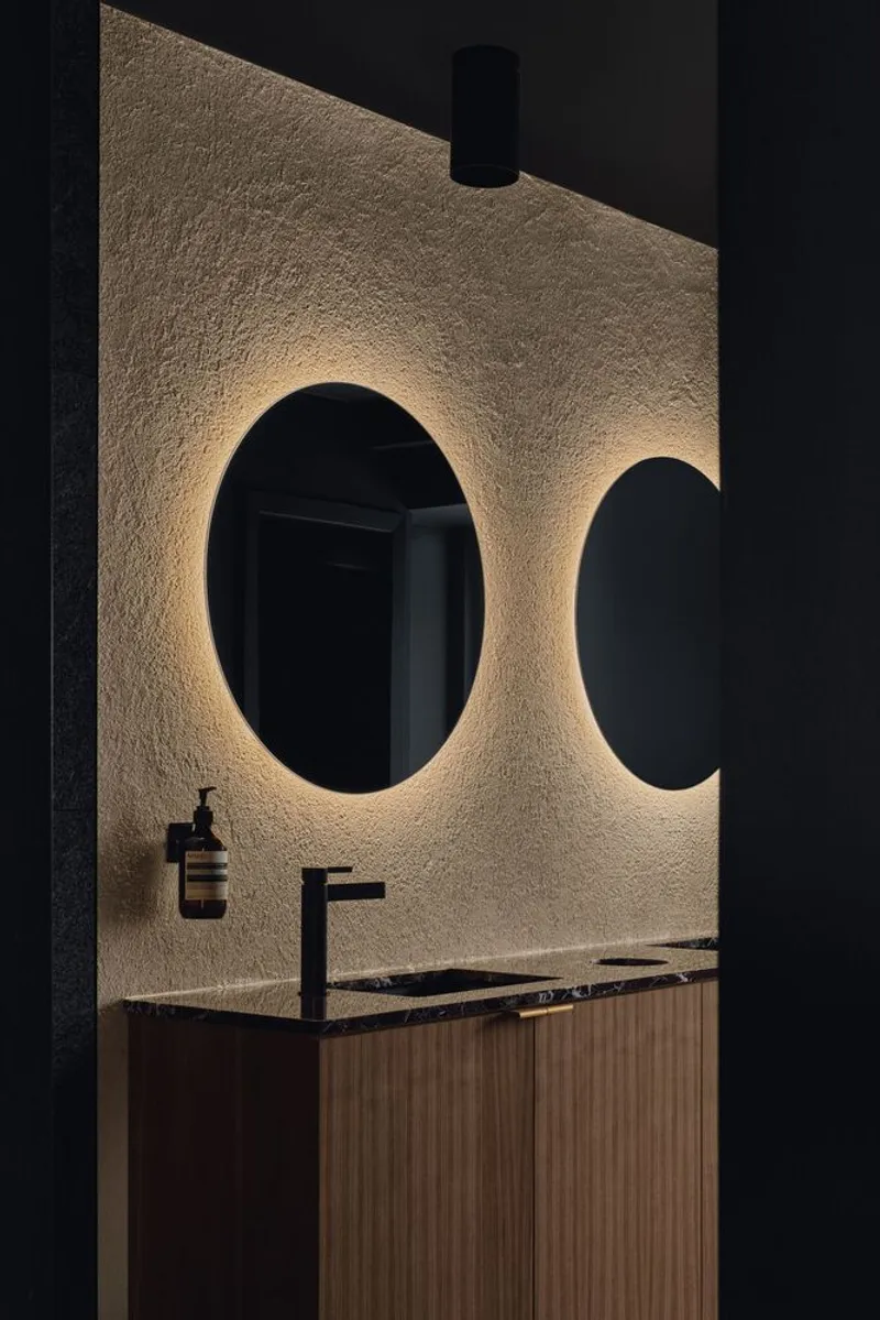Restroom vanity with backlit circular mirrors set into textured plaster wall at night