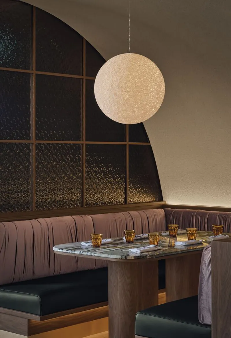 Spherical pendant light suspended above a marble-topped table with pink banquette seating and dark textured wall panels
