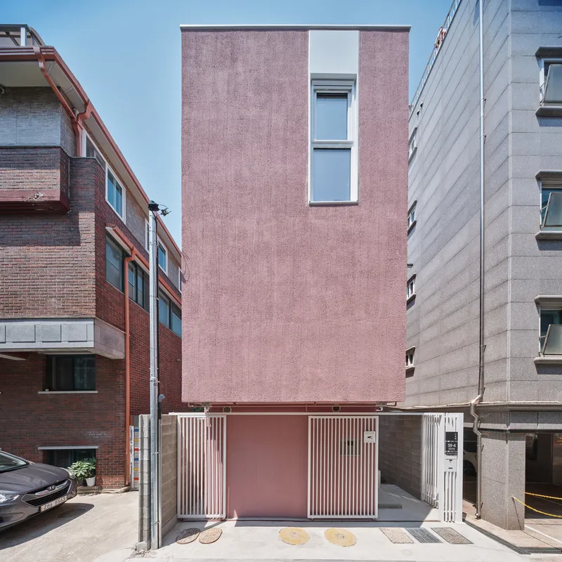 Front facade: four-storey dusty pink rendered tower with narrow vertical windows, concrete base, garage door, Korean alley