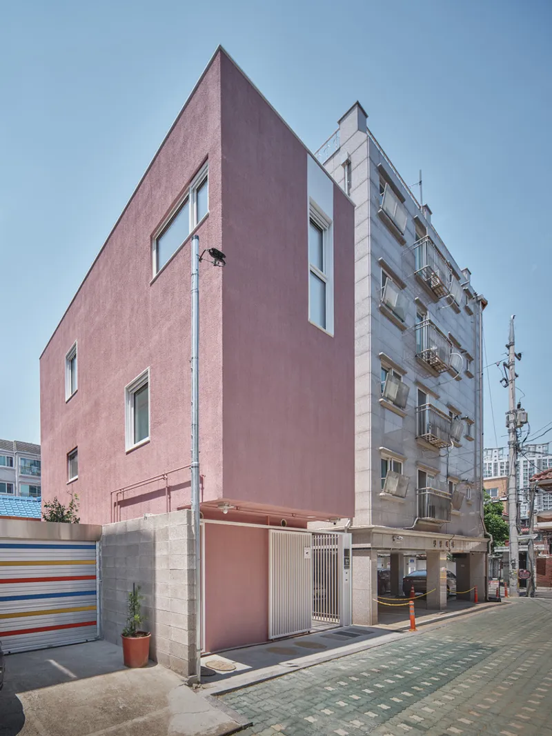 Three-quarter view: pink tower with angular top, concrete base, narrow alley, neighbouring apartment buildings