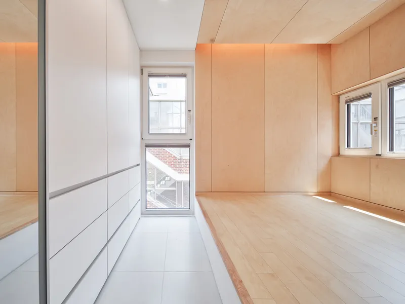 Plywood room: warm birch panel walls and ceiling, recessed LED strip lighting, white built-in storage, glass door to small balcony