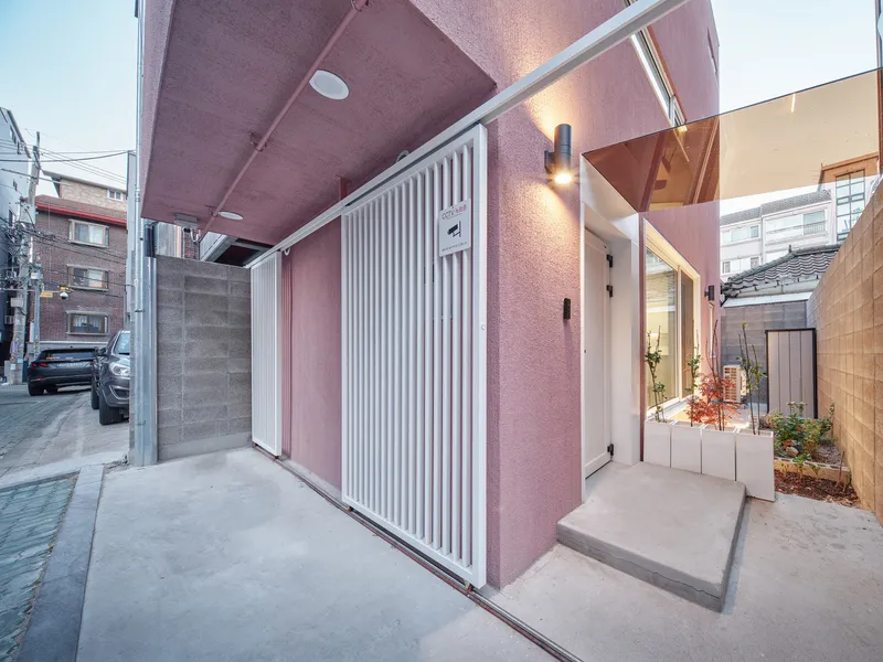 Entry: pink rendered soffit overhead, white slatted gate, small planted garden at the side, warm evening light