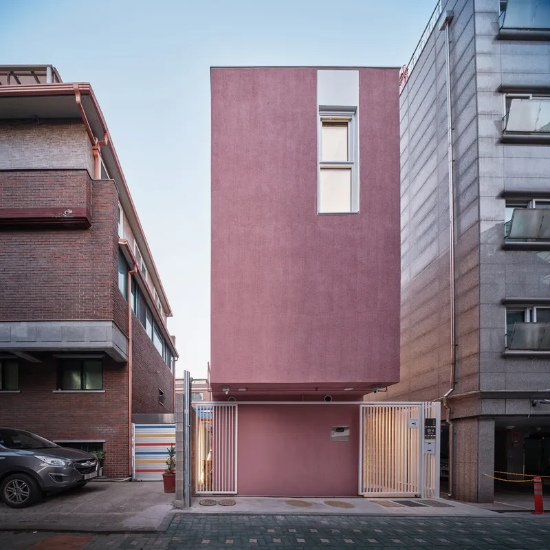 Dusk: pink facade with warm glow from interior, concrete base, timber door, car parked in the lane