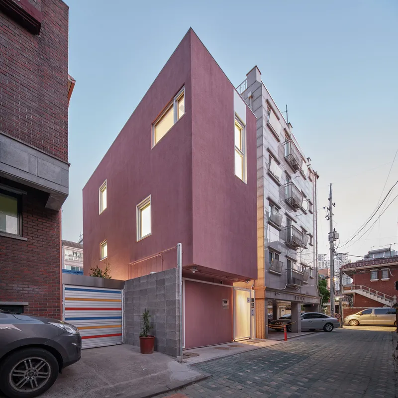 Dusk three-quarter: pink tower with lit windows, concrete block base, colourful striped element at ground level, neighbours behind