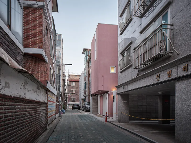 Alley context: pink house between brick and grey apartment buildings, Korean signage visible, dense urban neighbourhood